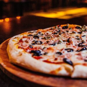 Close-up of a vegetable pizza with olives on a wooden board in a cozy restaurant setting.